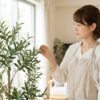 明るいリビングで、本物そっくりの人工観葉植物（オリーブの木）の葉に触れ、その質感を確かめる日本人女性。