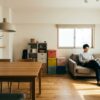 日本のリビングルームで、異なる家具ブランド（ニトリ、無印良品、イケアをイメージ）を組み合わせて快適に過ごす日本人男性。低価格で実用的な家具と、北欧デザインの小物が調和している。