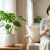 明るく清潔な日本人のリビングで、モンステラ、サンスベリア、ピレアなどの観葉植物が白い鉢に植えられ、木製のテーブルやスタンドに置かれている。背景で女性がソファーでガーデニング本を読みながら微笑む、虫がいない快適なグリーン空間。