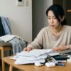 散らかった部屋のテーブルで、山積みの書類や物に囲まれ、疲れた表情で考え込む日本人女性。部屋の状態が心の余裕のなさを表している様子。