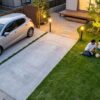 日本人の家族が暮らす住宅の庭で、駐車スペースのコンクリート（タマリュウ目地入り）と、主庭の天然芝、アプローチの砂利が美しくゾーニングされている。夕暮れ時に温かい照明が灯り、機能性と美観を両立している。