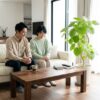 深みのある色合いのウォールナット材のテーブルと、隣に置かれた鮮やかなグリーンの観葉植物（ウンベラータ）。日本人の女性がテーブルにつき、植物を見ながらリラックスしている、明るい抜け感のあるモダンなリビングルームの風景。